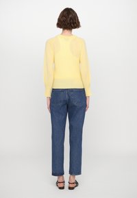 Gelbes Strickpullover mit figurbetonter Silhouette, gerippten Bündchen und Saum. Dazu hoch taillierte blaue Jeans und flache Sandalen.