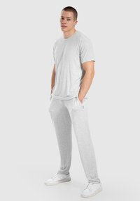 Jongeman staat met de handen in de zakken, draagt een lichtgrijs T-shirt, bijpassende joggingsbroek en witte sneakers op een effen achtergrond.