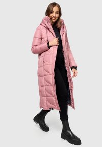 Long pink padded coat with a hood, featuring stitched quilted pattern and side pockets, paired with black ankle boots and pants.