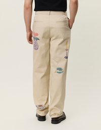 Μπεζ παντελόνι με πίσω κέντημα που αναπαριστά ένα βάζο, μία βάση κεριών και διάφορα αντικείμενα. Φτιαγμένο από βαμβάκι, διαθέτει χαλαρή γραμμή και πλευρικές τσέπες.