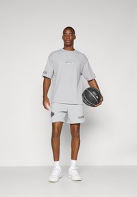 Šedé sportovní tričko s krátkým rukávem a šortkami s logem a nápisem Lakers. Má volný střih. Model drží basketbal.