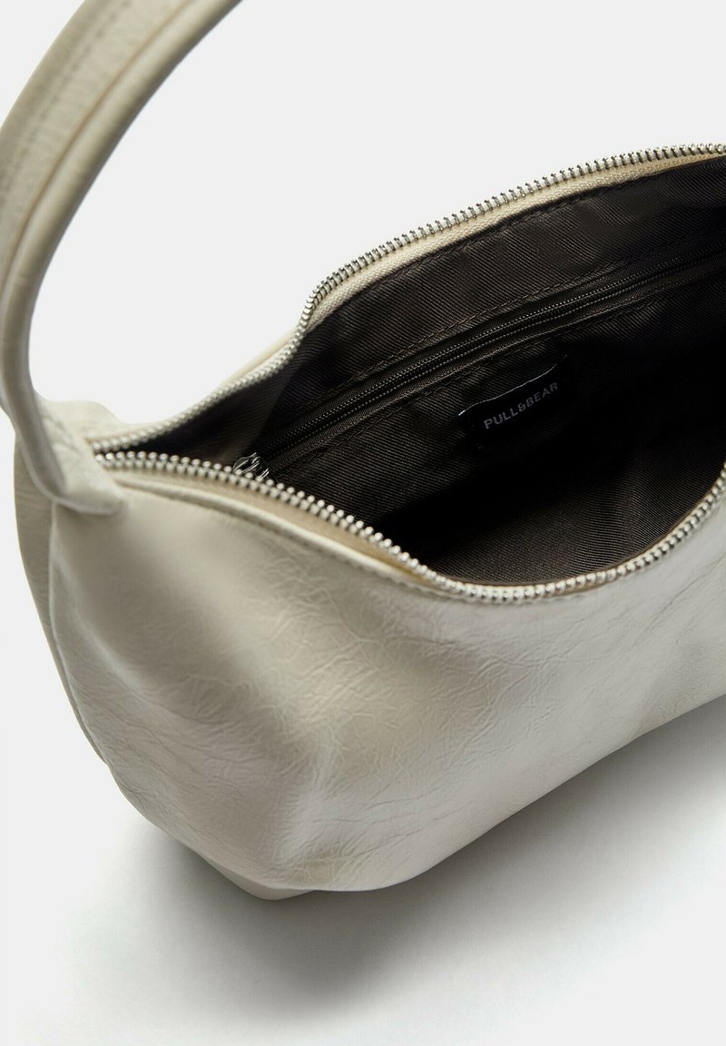 Bolso de mano blanco texturizado con cierre de cremallera. El interior es de color marrón oscuro y cuenta con un bolsillo adicional con cremallera. Tiene una sola correa para el hombro, curva.