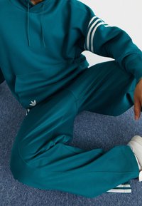 Teal hoodie med känguruficka, har tre vita ränder på ärmarna. Matchande teal joggingbyxor med slät, mjuk textur.