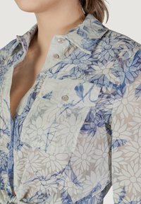 Transparante overhemdbloes met een bloemenpatroon in blauw en wit, voorzien van een borstzak en een gladde textuur.