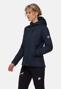 Blond kvinna i en marinblå huva jacka och svarta byxor med loggor, står på sidan med en hand i jackfickan.