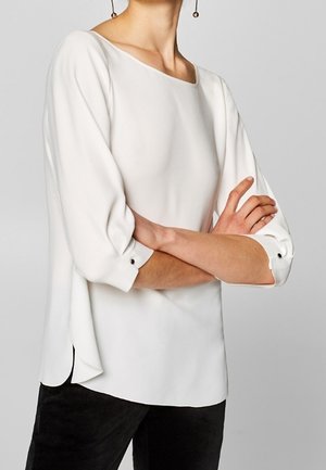 Nainen pukeutuneena pitkähihainen valkoinen paita, jonka hihat on kääritty, ja mustat housut, kädet ristissä vyötäröllä harmaata taustaa vasten.