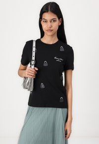 Melnas īsām piedurknēm tees ar izšūtiem pelēkiem maisiņiem un tekstu. Modelis tur sudraba plecu somu. Zemē viegla zaļa svārku plisē.