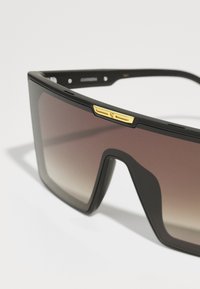 Occhiali da sole neri oversize con lenti marroni sfumate. Presentano un accento dorato sulla montatura e una finitura opaca. Design angolare.