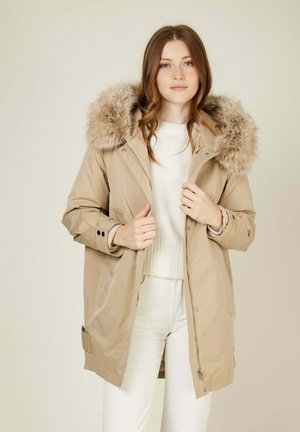 Parka beige avec une capuche en fourrure, manches longues, fermetures éclair et boutons pression, poches latérales, portée sur un pull blanc et un pantalon clair.