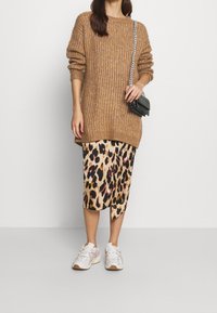 Donna che indossa un maglione spesso marrone, una gonna midi con stampa leopardata, scarpe da ginnastica bianche e una piccola borsa a tracolla nera con una catena argentata.