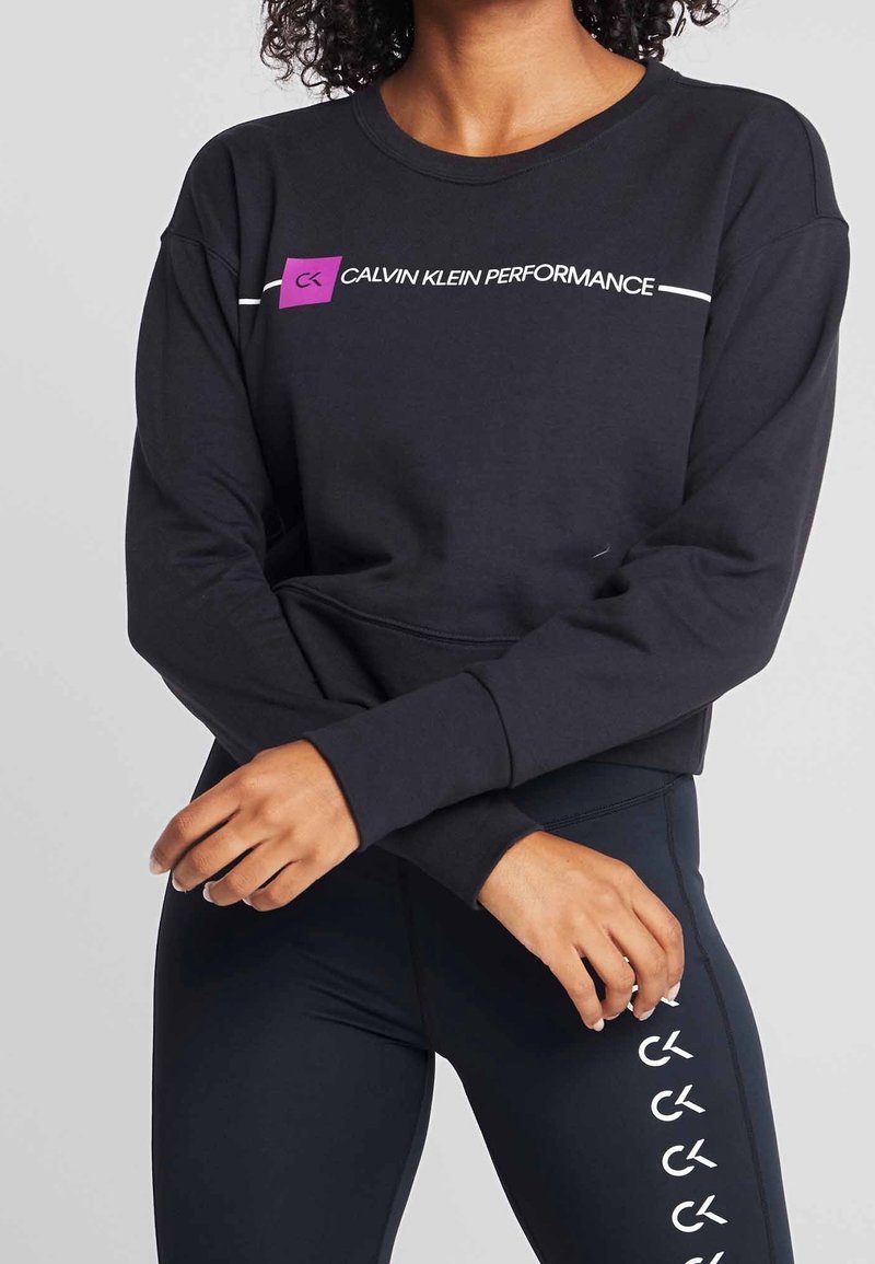 Persoon in een zwart Calvin Klein Performance langekoope top en zwarte leggings met herhaalde CK-logo's op de zijkant.