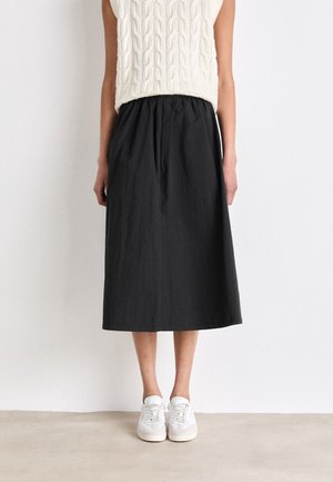 Woman wearing a sleeveless white knit top, black midi skirt, and white sneakers standing against a plain white wall.