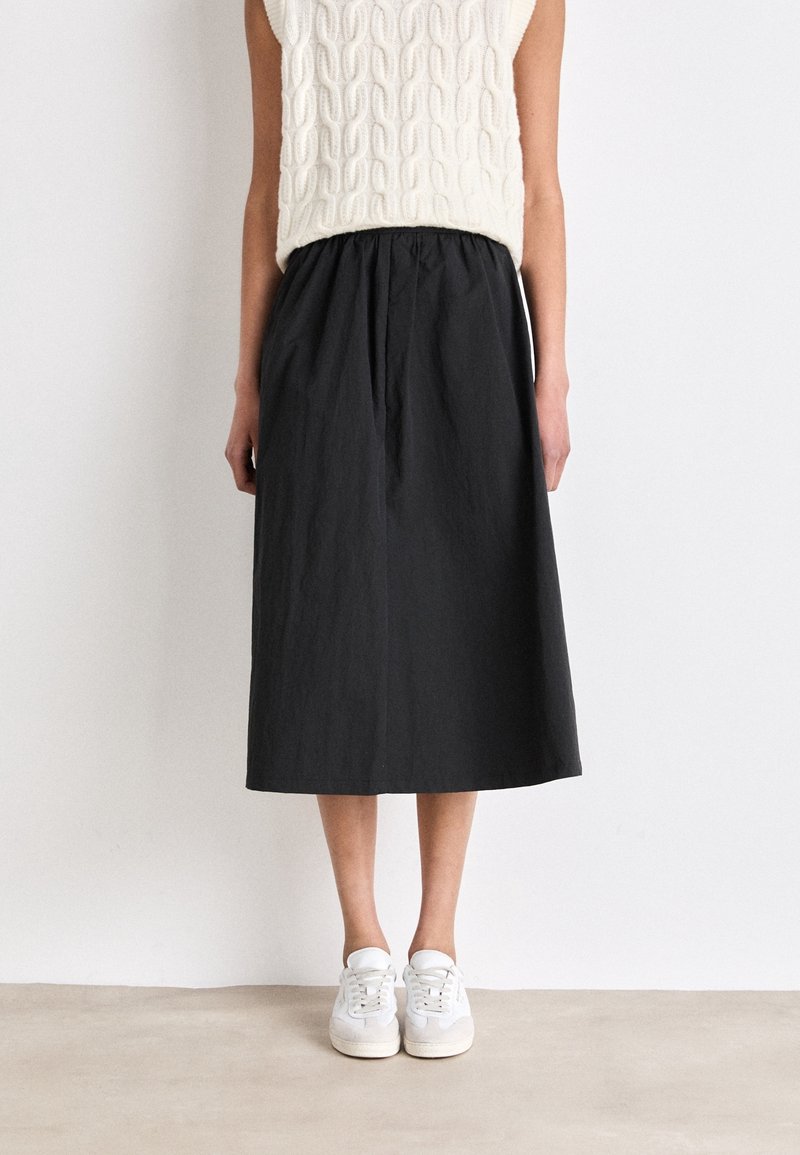 Femme portant un haut blanc sans manches en maille, une jupe midi noire et des baskets blanches, debout contre un mur blanc uni.
