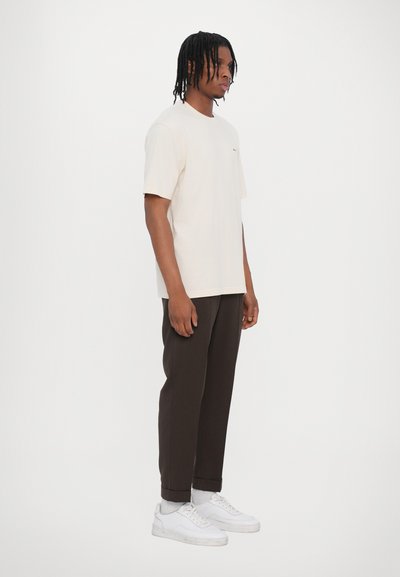 Camiseta de algodón beige con mangas cortas, combinada con pantalones oscuros marrones y zapatillas blancas. Diseño sencillo con un corte relajado.