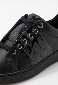 Černé kožené tenisky s lesklou vzorovanou částí, černými šňůrkami provlečenými stříbrnými průchodkami a texturovanou gumovou podrážkou.