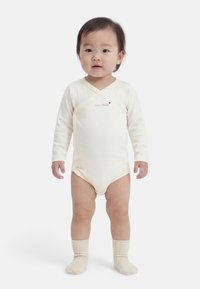 Νήπιο όρθιο που φορά ένα μπεζ ολόσωμο ρούχο με μακριά μανίκια και το κείμενο «mon trésor», μαζί με μπεζ καλτσάκια, σε λευκό φόντο.