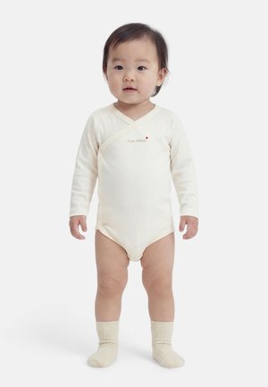 Tout-petit debout portant un body beige à manches longues avec le texte "mon trésor" et des chaussettes beige assorties sur un fond blanc.