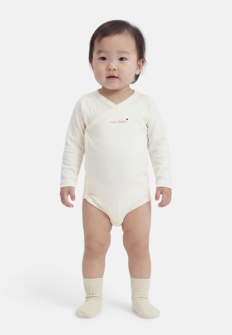 Νήπιο όρθιο που φορά ένα μπεζ ολόσωμο ρούχο με μακριά μανίκια και το κείμενο «mon trésor», μαζί με μπεζ καλτσάκια, σε λευκό φόντο.