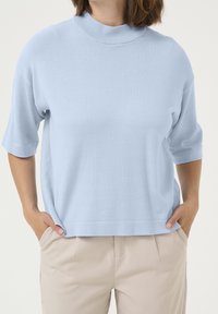 Ljusblå stickad topp med kort ärm och hög halsringning, i avslappnad passform, med ribbade ärmslut och fåll. Toppad med beiga byxor.