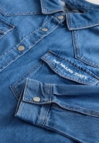 Denim jacket featuring a blue hue, snap buttons, and a chest pocket with "THE ROLLING STONES" embroidered in white. Textured fabric and stitching details.