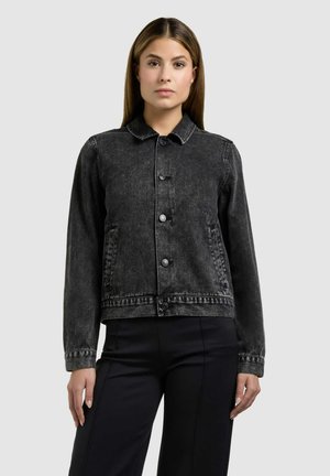 Žena s rovnými hnědými vlasy, oblečená v zapnuté tmavě šedé džínové bundě a černých kalhotách, stojící před prostým světlým pozadím.