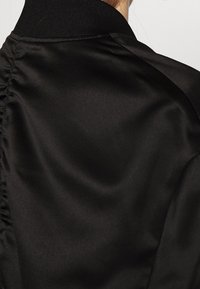 Zwarte satijnen bomberjack met een ribbelkraag, een soepele textuur en gathered stiksels langs de naad.