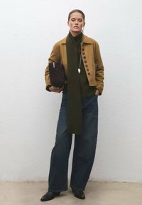 Giacca in camoscio marrone con bottoni, sciarpa lavorata a maglia verde scuro, pantaloni ampi in denim scuro e stivaletti neri, con in mano una borsa marrone.