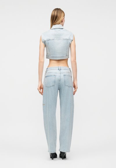 Γυναίκα που φοράει ανοιχτό γαλάζιο κοντό αμάνικο τζιν μπουφάν και τζιν παντελόνι με φαρδιά μπατζάκια και ψηλοτάκουνες μαύρες γόβες, στέκεται γυρισμένη με την πλάτη.