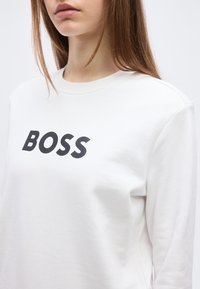 Νεαρός με μακριά μαλλιά που φοράει λευκό φούτερ με τη λέξη "BOSS" τυπωμένη με έντονα μαύρα γράμματα στο στήθος.