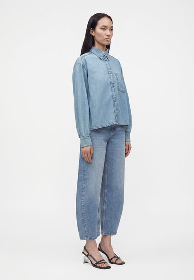 Femme debout portant une chemise en jean bleu clair, un jean taille haute à jambes larges et des sandales à talons noires à bout ouvert sur un fond uni.