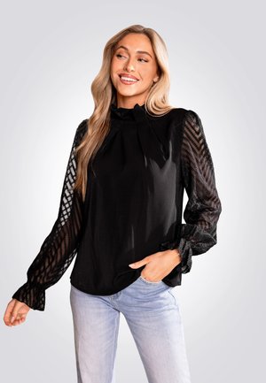 Zwarte blouse met getextureerde, doorschijnende mouwen met een zigzagpatroon, hoge hals en ingezette manchet. G draag in combinatie met lichtblauwe jeans.