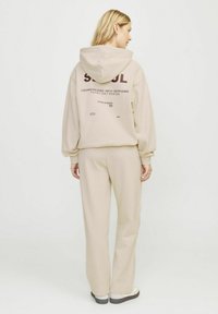 La sudadera beige y los pantalones a juego presentan un gráfico en la parte posterior con "SEOUL" y texto adicional. Tejido suave con un corte relajado y puños acanalados.