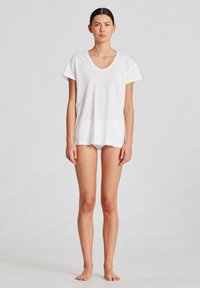 Tricou alb din bumbac, cu decolteu în V, mâneci scurte și croială lejeră. Modelul stă desculț, purtând slipuri albe asortate.