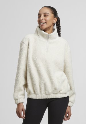 Kvinde med flettet hår iført en hvid fleecepullover med halv lynlås og sorte bukser, smilende og kigger til siden.