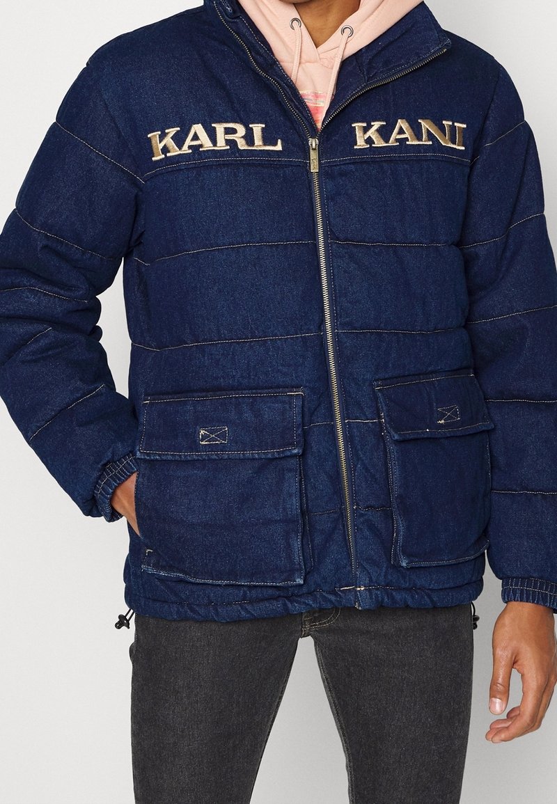 Homme portant une veste matelassée bleu marine avec fermeture éclair à l'avant, grandes poches à rabat, et "KARL KANI" brodé sur la poitrine, sur un sweat à capuche rose.