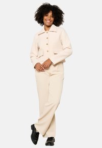 Izgriezta bēša jakas ar zelta pogām un divām priekšējām kabatām, kombinēta ar bēšu platām biksēm un melniem lakotiem apaviem.