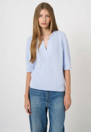 Young woman with long straight hair wearing a light blue knit top with a deep V-neck and blue jeans against a plain background.