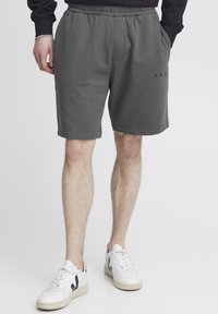 Grå bomuldsshorts med elastisk talje, sidelommer og et logo på venstre ben. Bæres med hvide sneakers.