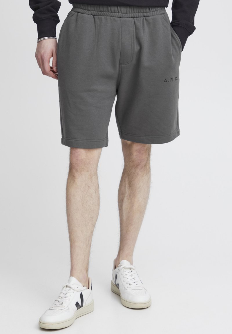 Grå bomuldsshorts med elastisk talje, sidelommer og et logo på venstre ben. Bæres med hvide sneakers.