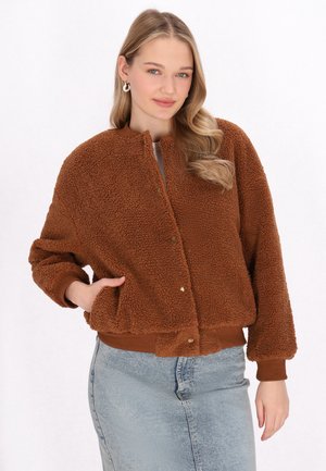Jachetă bomber din blană maro, pufoasă, cu guler rotund, nasturi tip snap și manșete ribdate; purtată cu o fustă din denim albastru deschis.