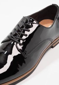 Chaussure habillée en cuir verni noir avec fermeture à lacets, bord de semelle marron et coutures visibles sur fond blanc.
