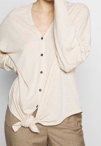 Ljusbeige cardigan med V-ringning, knappstängning och långa ärmar, med en knuten design vid midjan. Slät textur.