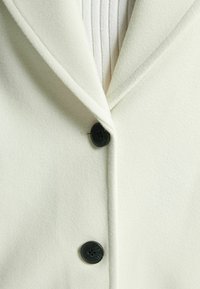 Kremfärgad ullkappa med stuck kollär och tre svarta knappar. Mjuk textur med en slät yta. Närbild av framsidan.