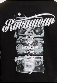 Svart t-skjorte med et grafisk design av en boombox i grått, med teksten "Rocawear" og en setning under i hvitt.