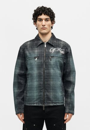 Joven con el pelo rizado y corto lleva una chaqueta de cuadros negra y gris descolorida con cremallera, dos bolsillos con cremallera en el pecho y el texto "Off-White" en el pecho.