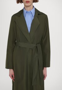Olijf groene trenchcoat met een riem, gekartelde kraag en lange mouwen, gelaagd over een lichtblauw overhemd met knopen. Mals stof textuur.