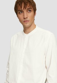 Camisa blanca de botones con cuello de banda, hecha de tela texturizada, que presenta un sutil logo bordado en el pecho y botones visibles.