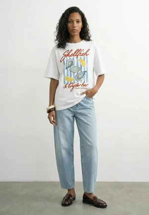 Kvinde med krøllet hår iført en hvid t-shirt med skaldyrsmotiv, lyseblå jeans, armbånd og loafers med leopardprint, stående foran en ensfarvet baggrund.