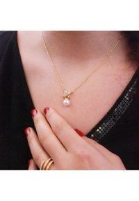 Gouden halsketting met een delicate ketting, voorzien van een ronde parelpendant en een bladerenaccent boven de parel. Zwart top met zichtbare pailletten.