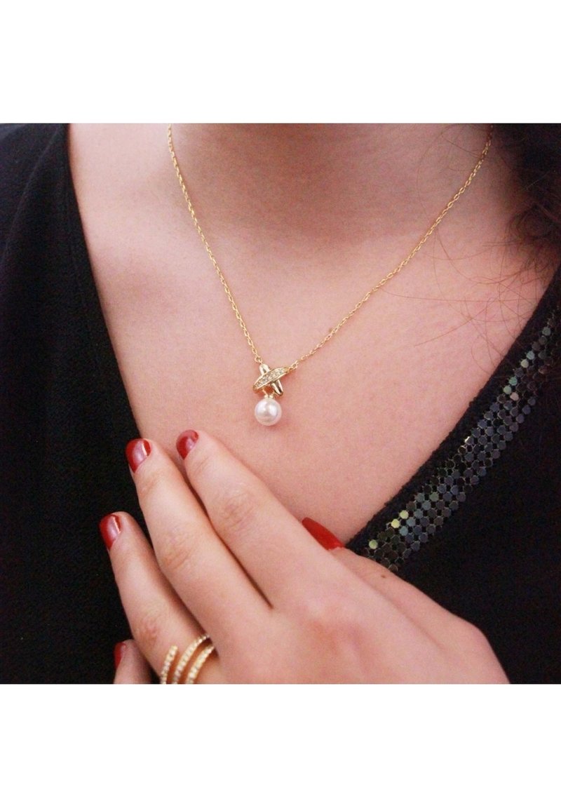 Gouden halsketting met een delicate ketting, voorzien van een ronde parelpendant en een bladerenaccent boven de parel. Zwart top met zichtbare pailletten.